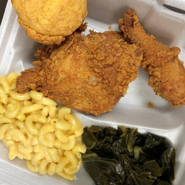 Fried chicken, cornbread, macaroni and cheese, and collard greens in a white takeout container.