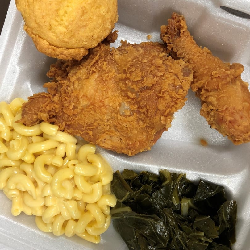 Fried chicken, cornbread, macaroni and cheese, and collard greens in a white takeout container.